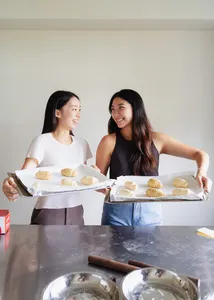 Two learners holding trays of shio pan buns while smiling at the counter, with rolled dough and bowls nearby during workshops in Singapore