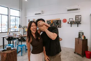 Laughing couple posing in a bright silversmithing studio with workbenches and tools behind them at Fat Anvil Studios, Singapore