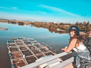 Helmed traveller smiling at sunrise on a riverside bridge in Hoi An, with long fishing boats moored in calm water