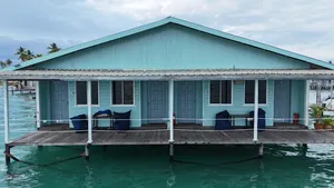 Banglo air kayu berwarna hijau kebiruan di atas tiang di Semporna, dengan tempat duduk di anjung hadapan dan laluan pejalan kaki di atas laut yang tenang