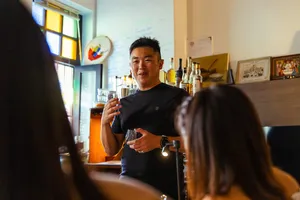 Host explaining flavours during a small-batch whisky tasting in Singapore, reaching up a bottle as guests listen in a home bar