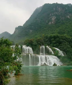 Tiered waterfalls flowing into a green river with mist, backed by steep forested mountains under an overcast sky