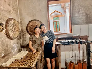 Two women smiling in a Hanoi tofu-making kitchen beside bamboo mats of pressed tofu, with doorway light behind them