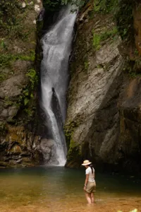 Traveller in a cap standing near a tall waterfall pouring into a rocky pool during a Borneo Overland wild Sabah road trip