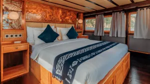 Warm-toned deluxe phinisi bedroom with a neatly made double bed, curtained windows, and sea views during a 4D3N shared Komodo cruise, Komodo Island