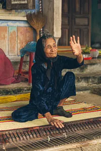 Local artisan seated and waving while weaving on a patterned floor mat in Hoi An, showcasing an old-town craft at dawn