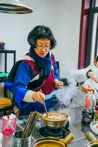 Fancy stirring a pot on a stovetop while cooking Yunnan food in a Lijiang kitchen, with herbs and sauces on the table