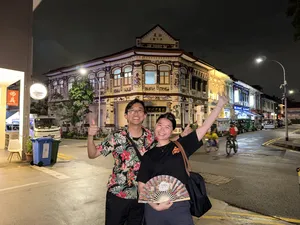 Two people posing on a night street in Geylang, Singapore, holding a folding fan in front of a heritage shophouse