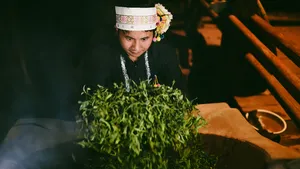 Seorang perempuan setempat dengan hiasan kepala bordir memegang seikat herba hijau segar di ambang pintu desa di Xishuangbanna