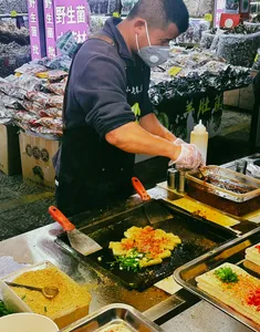 Local cook at a Lijiang street-food stall spreading savoury batter on a sizzling griddle with chopped greens and sauce