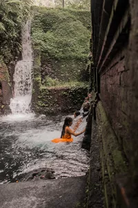 Yoga participant in a narrow waterfall stream in Indonesia, seated on an orange mat with arms raised in a practise pose