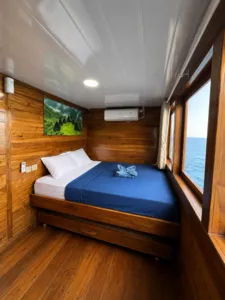 Wooden phinisi cabin with air-conditioned double bed, curtain-side window and wall art on a shared Komodo cruise from Labuan Bajo