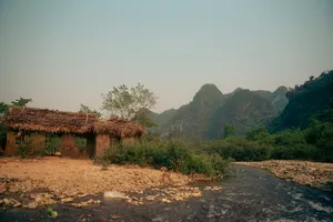 Thatched-roof stilt hut by a rocky stream with hazy limestone mountains in Phong Nha on a local-tribe tour