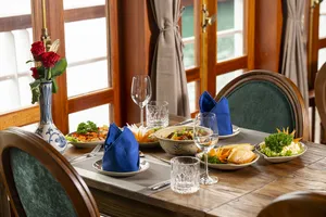 Set dining table aboard Cat Ba Ocean Cruises with bowls of food, red flowers, and blue napkins in cosy wooden seating