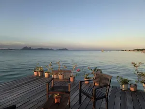 Tempat duduk kayu di dek banglo atas air di Semporna pada waktu senja, dengan tanaman pasu, laut yang tenang, dan pulau-pulau di kejauhan