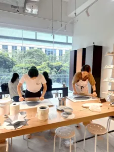 Para peserta di meja kerja tembikar membentuk hasil seramik di sebuah studio cerah di Singapura, dengan peralatan tersusun dan tingkap besar di belakang