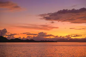 Sunset over calm sea with dark island silhouettes on the horizon, warm orange clouds reflecting on the water at dusk