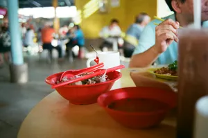 Red hawker bowls on a dining table in Singapore, with diners’ hands enjoying noodles and side dishes together