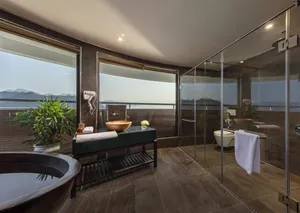 Glass-walled shower and bathtub in an Oasis Bay Party Cruise suite, with windows overlooking Halong Bay scenery