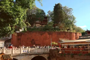 Visitors on a riverside balustrade beside a calm canal and carved red wall in Lijiang Old Town with a local guide