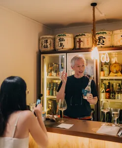 SakeMama pouring rare sake for guests during a Singapore tasting in a cosy shop, with glowing bottles and jars behind