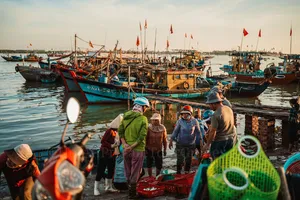 Small group of helmed travellers walking along a Hoi An harbour dock at sunrise, surrounded by fishing boats with flags overhead