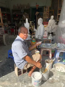 Elderly artisan painting small figurines and surrounded by white Buddha sculptures at a craft shop in Ho Chi Minh City
