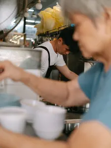 Chef preparing food at a kitchen counter, with diners blurred nearby, on a Joo Chiat food tour in Singapore