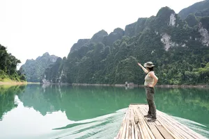 Solo traveller on a wooden raft deck with a straw hat rests by the calm green water and limestone cliffs at Cheow Lan Lake