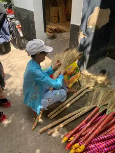 Saigon artisan in a straw hat weaving and packing broom brushes, with rows of finished thatch brooms laid out nearby