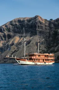 Sailing phinisi passing rugged volcanic hills in calm blue water, for a 3-day shared Komodo cruise from Labuan Bajo