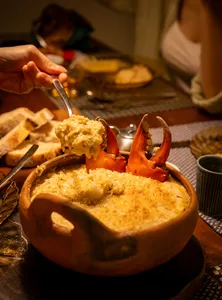 Hand lifting orange prawns from a clay bowl of creamy stew, with bread on the side during a 5-course Patagonian-inspired dinner in Singapore