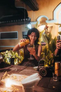 在会安温暖的灯笼灯光下，一位女子在热闹的品鉴桌旁把罐装柑橘鸡尾酒倒入杯中，周围的人举着手机记录这一刻
