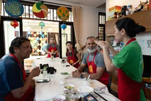会安烹饪课上，系着红围裙的客人在餐桌旁准备食材，主人在彩灯旁讲解指导