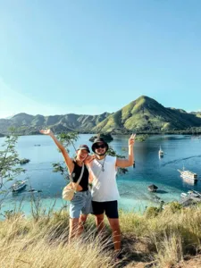 Two travellers posing with arms up on a grassy coast viewpoint overlooking a bay with anchored boats on a shared Komodo cruise — Komodo Island