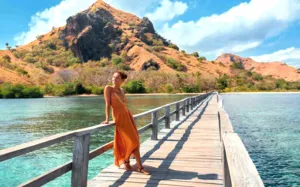 Traveller in a rust-coloured dress walking along a wooden pier over clear water with rugged hills in the distance, in Labuan Bajo