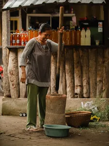 A man prepares drinks at an outdoor village counter in Lombok, with rows of amber bottles and a serving cart behind him