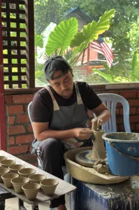 Ceramic artist at Ilham Ceramic in Langkawi works on a pottery wheel, with finished cups on a tray beside a sunlit window.