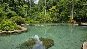 People relax and wade in shallow turquoise Blue Spring water next to floating rafts, surrounded by rainforest in Khao Sok National Park