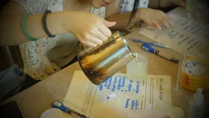 Hand pouring wax into a small candle jar on a Singapore worktable, with fragrance bottles and labels beside it in a perfume workshop