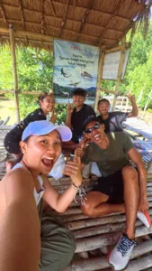 Sekumpulan pengembara tersenyum di ruang tempat duduk kayu berbumbung rumbia di sebelah papan tanda ilustrasi bakau di Labuan Bajo