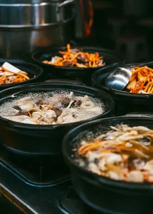 Rows of black soup pots at a Lijiang food stall with bubbling broth, fish pieces, and orange shredded vegetable topping
