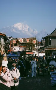 Evening street market in Lijiang with lantern-lit shopfronts, shoppers bundled up, and a snow-capped mountain towering behind