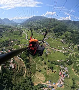 Tandem paragliding from behind with a helmeted rider guiding below the canopy over a valley of terraces and winding roads in Sapa