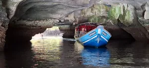 Small blue boat carrying passengers floats inside a rocky cave pool in Langkawi, with water ripples and sunlight at the opening