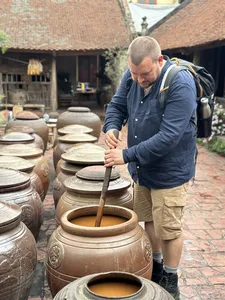 Visitor stirring liquid in a large clay jar among stacked pottery in a courtyard at Duong Lam ancient village near Hanoi during rice farming