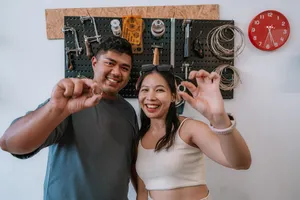 Smiling couple holding handmade rings in front of a wall packed with jewellery-making tools at Fat Anvil Studios, Singapore