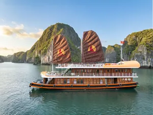 Cat Ba boutique cruise boat with tall sail covers cruising past limestone hills under a bright blue sky