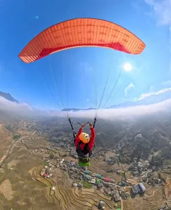 Paragliding rider in a red jacket reaching up beneath an orange canopy over terraced valley slopes, Sapa