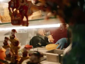 Person adding toppings behind a counter, viewed through reflections and motion blur in a Joo Chiat shop in Singapore